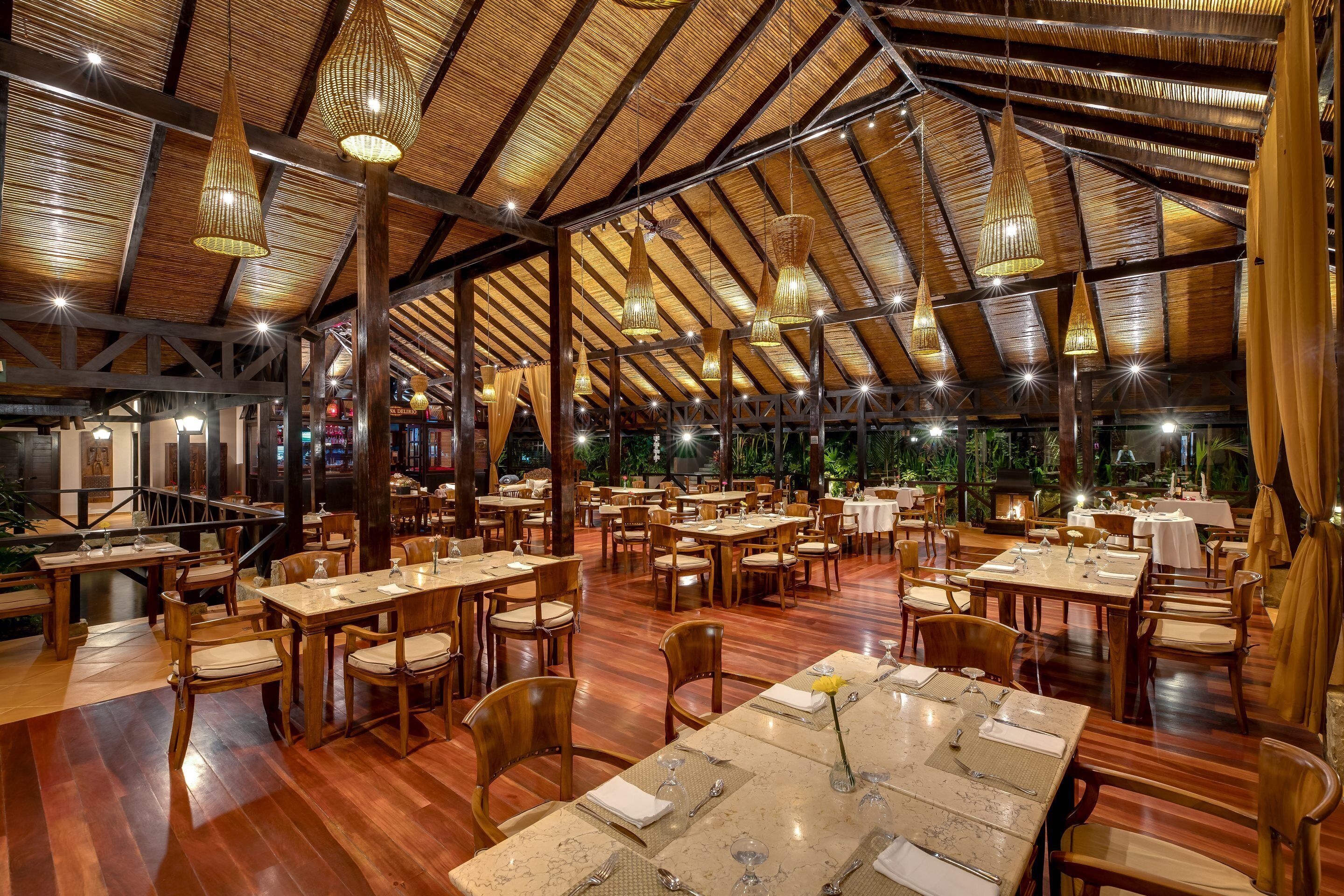A spacious restaurant with wooden tables and chairs, a high ceiling with hanging lights, and a view of the outdoors.