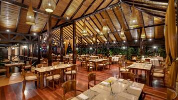 A spacious restaurant with wooden tables and chairs, a high ceiling with hanging lights, and a view of the outdoors.