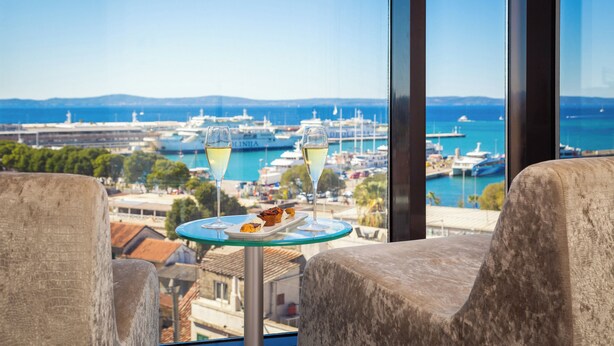 Un balcón con una mesa dispuesta para dos personas, con vistas a un puerto deportivo con barcos y un cielo azul despejado.