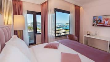 Habitación de hotel con una cama grande, un escritorio, una silla, un televisor y un balcón con vista al mar y barcos.