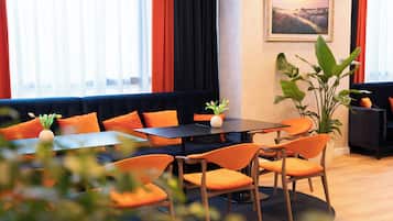 Un moderno vestíbulo de hotel con un área de estar que incluye sillas anaranjadas y una mesa negra, una gran planta en maceta y un cuadro con un paisaje en la pared.