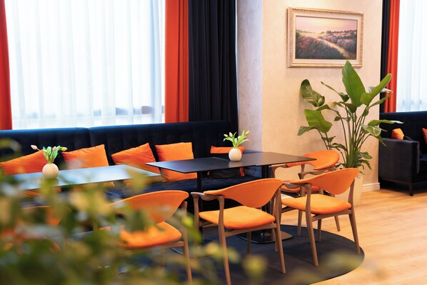 A modern hotel lobby with a seating area featuring orange chairs and a black table, a large potted plant, and a framed landscape painting on the wall.
