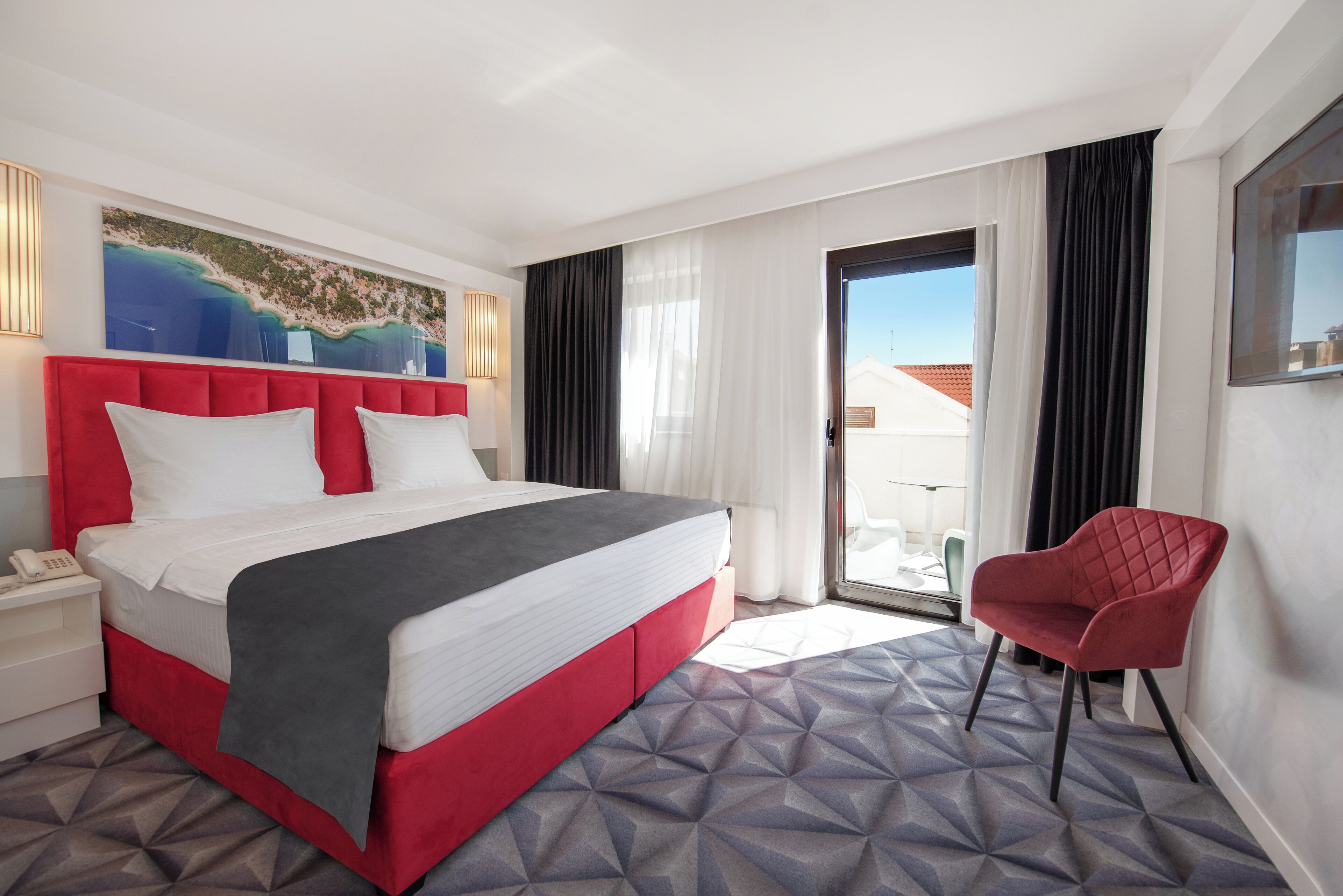 A hotel room with a large bed, a red chair, a painting of a beach, and a view of buildings through the window.