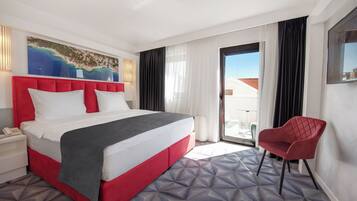 A hotel room with a large bed, a red chair, a painting of a beach, and a view of buildings through the window.