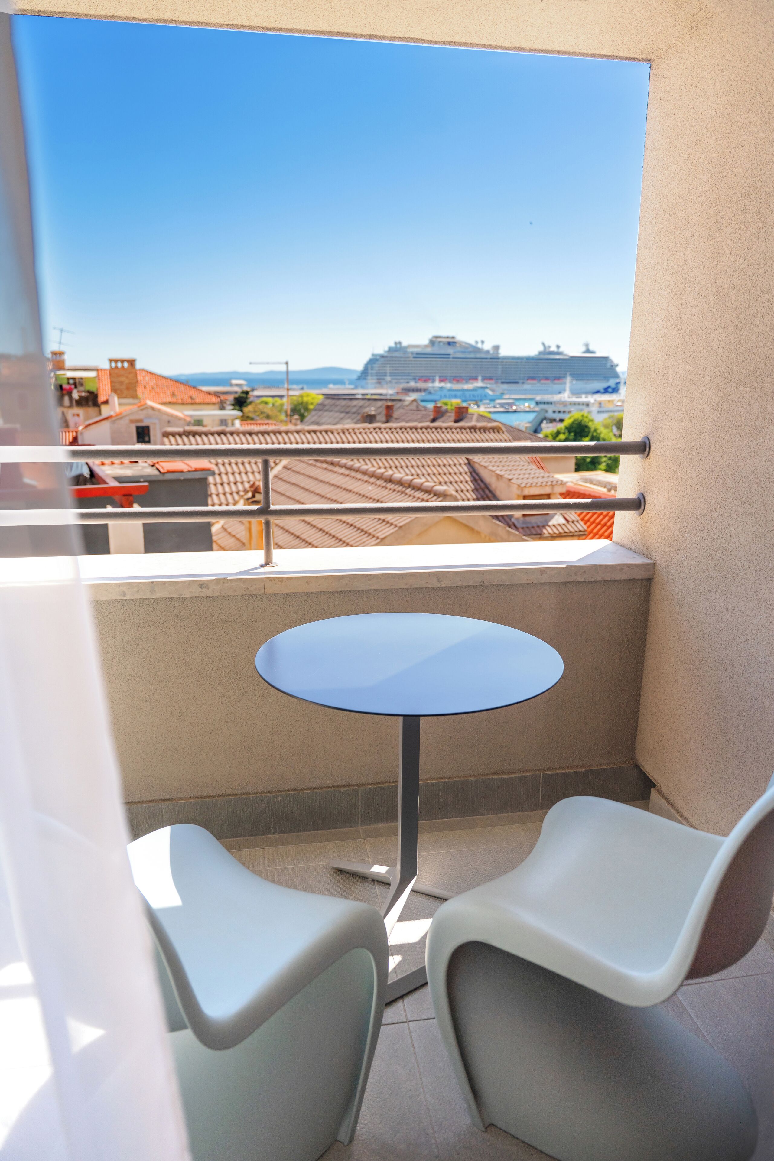 A balcony with a small round table and two white chairs, overlooking a cityscape with buildings and a large ship in the distance.