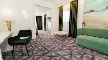 Ein Hotelzimmer mit einem geometrisch gemusterten Teppich, einem grünen Sofa, einem grünen Sessel, einem runden Tisch und einem Wandbild mit einer Strandansicht.