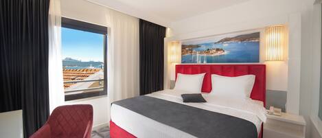 Une chambre d’hôtel avec un grand lit, une tête de lit rouge, une vue sur la mer, un fauteuil rouge et une table de chevet avec un téléphone.