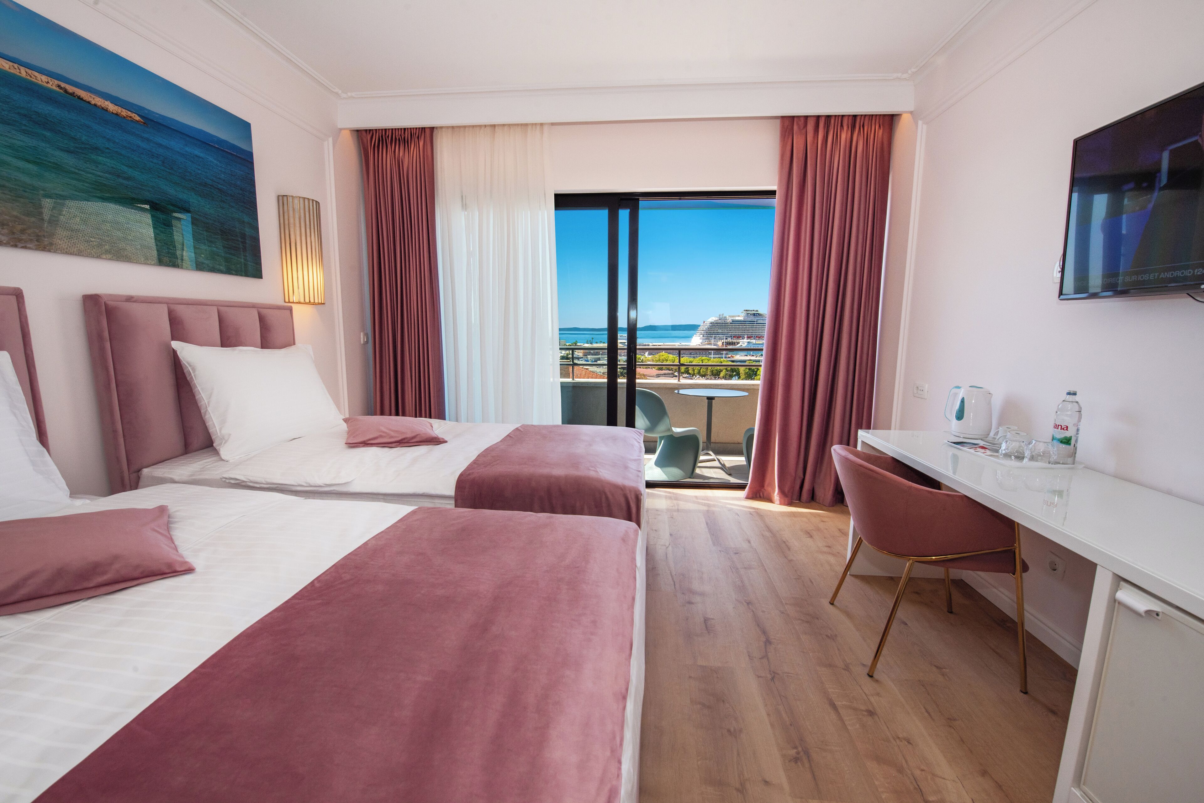 A hotel room with two beds, a desk, a chair, a TV, and a balcony with a view of the sea and buildings.