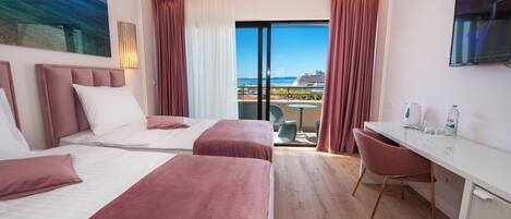 A hotel room with two beds, a desk, a chair, a TV, and a balcony with a view of the sea and buildings.