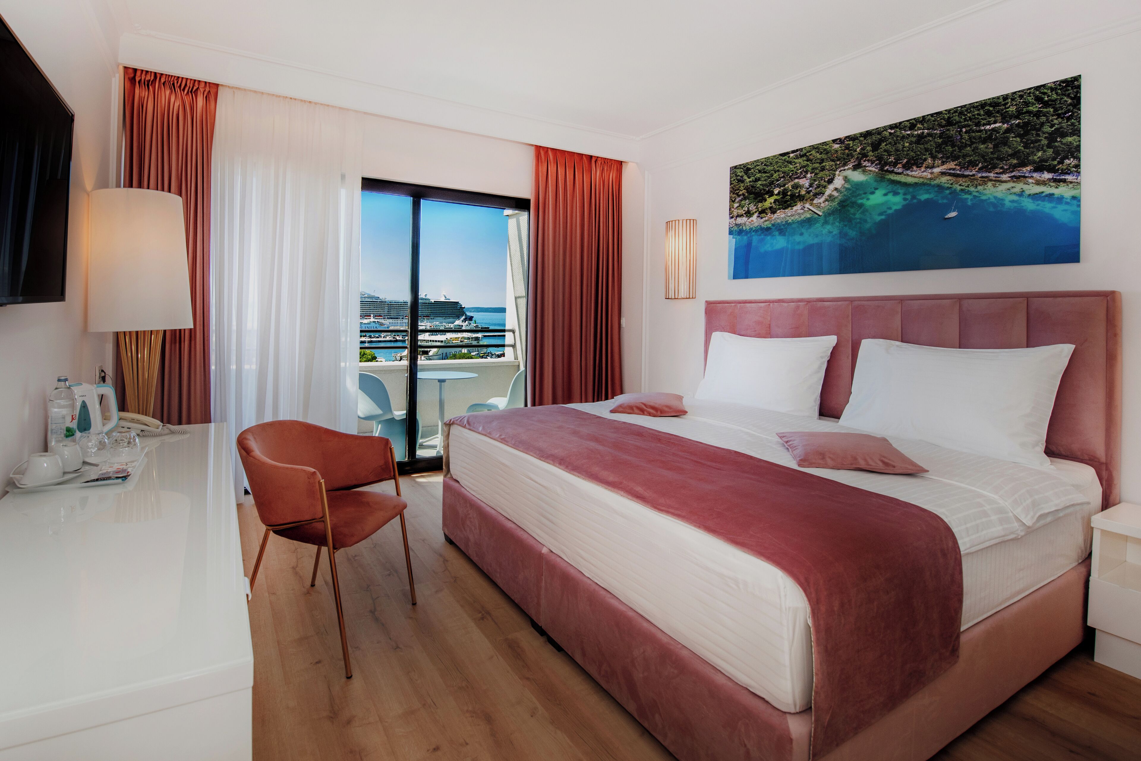 A hotel room with a large bed, a desk, a chair, and a view of a coastal area.
