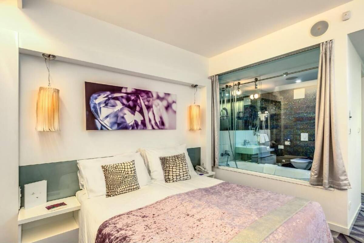Un dormitorio con cama, una mesita de noche, una lámpara de noche, una obra de arte en la pared, una ventana con vista a un baño y una cortina.