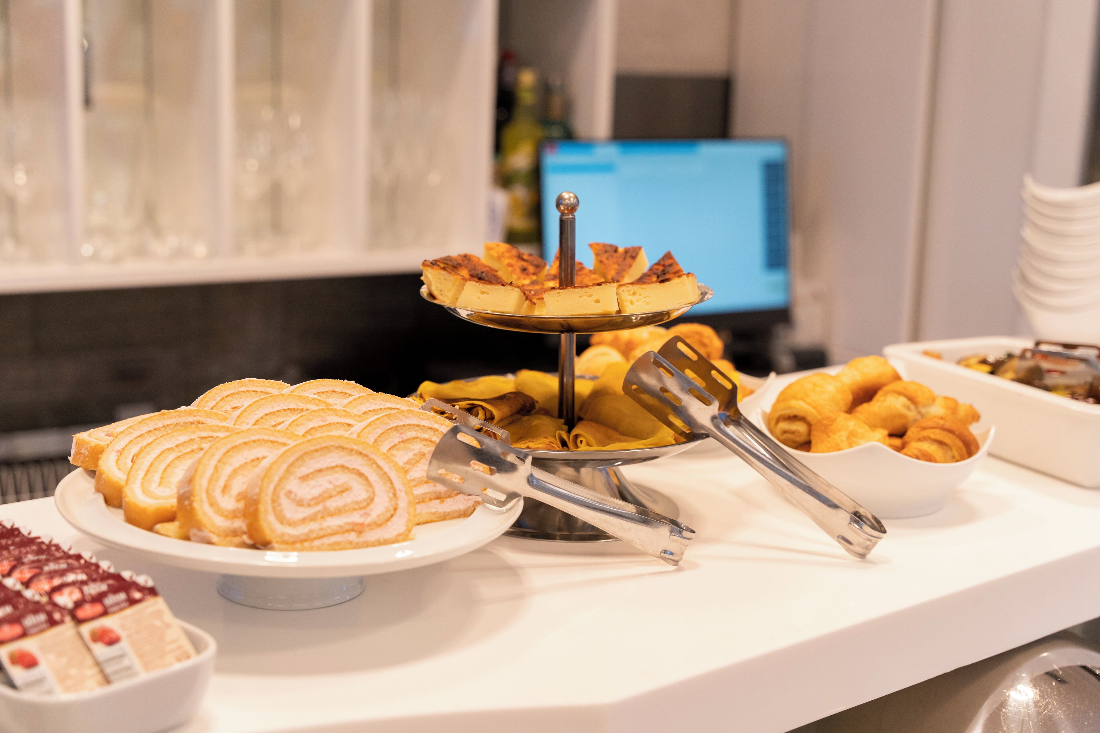 A buffet table with various pastries and desserts, including a cake on a stand, tongs, and bowls of food.