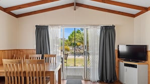 A room with a wooden dining table, chairs, a TV on a cabinet, and a sliding glass door leading to an outdoor area with a view of greenery. - Discovery Parks - Moama West (Moama)