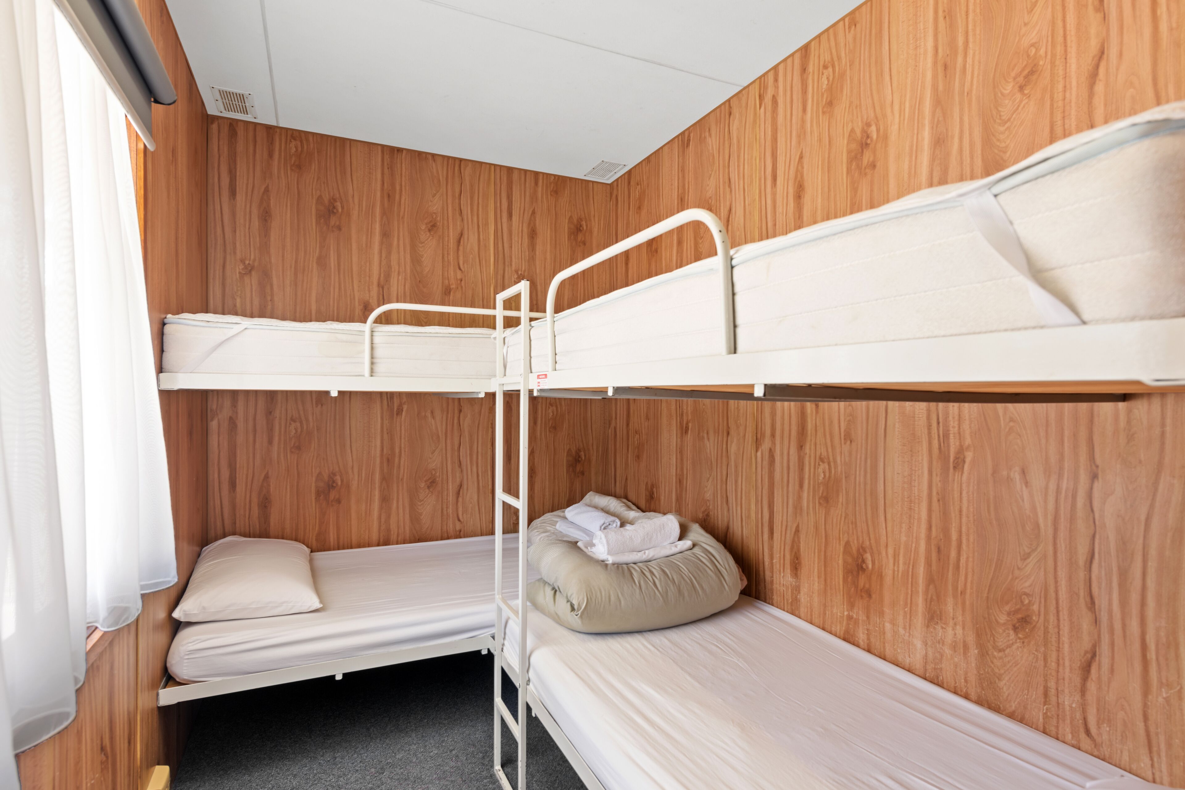 A bunk bed room with wooden paneling, a window with curtains, and a small table.