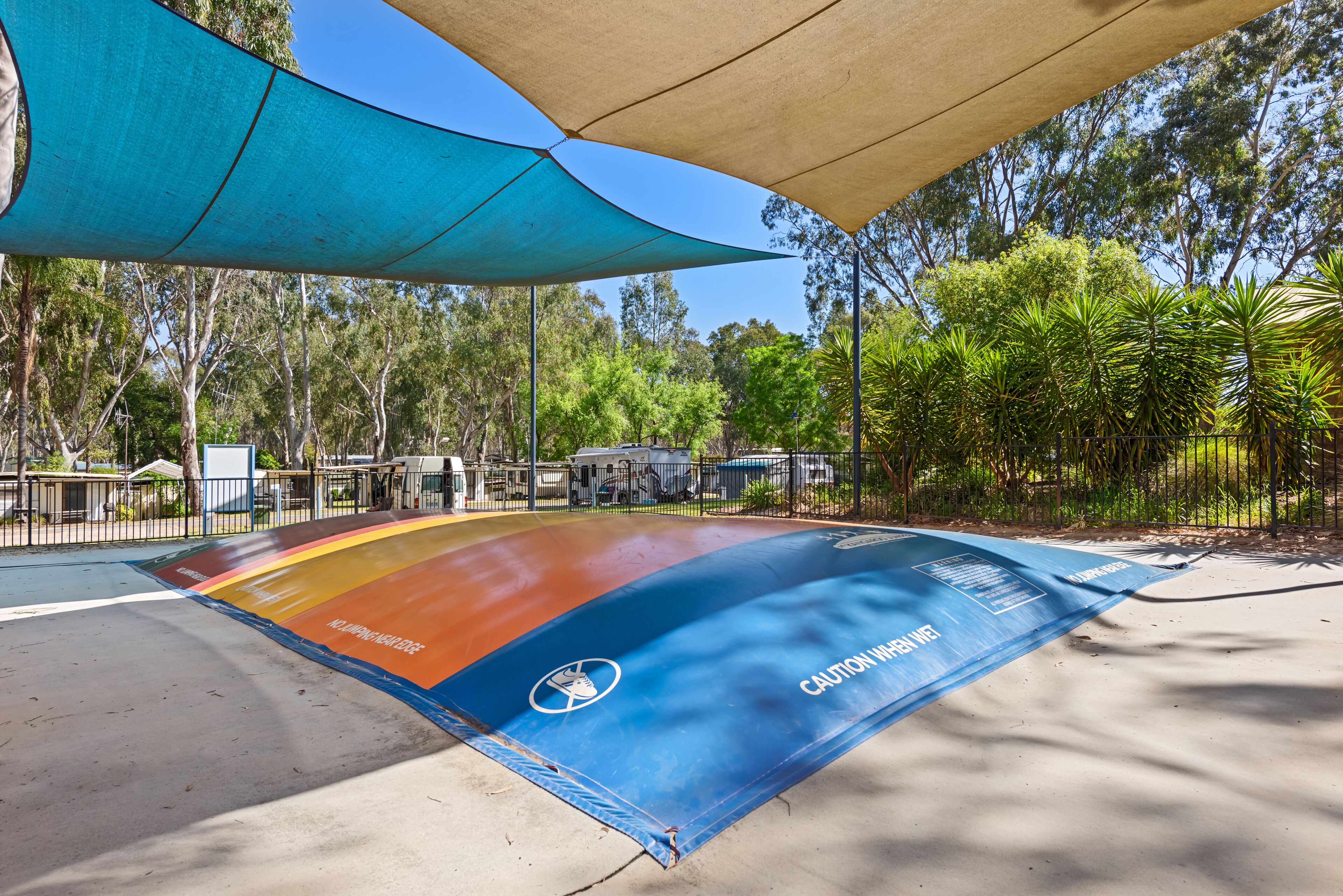 A shaded outdoor area with a colorful safety mat, trees, and a fence.