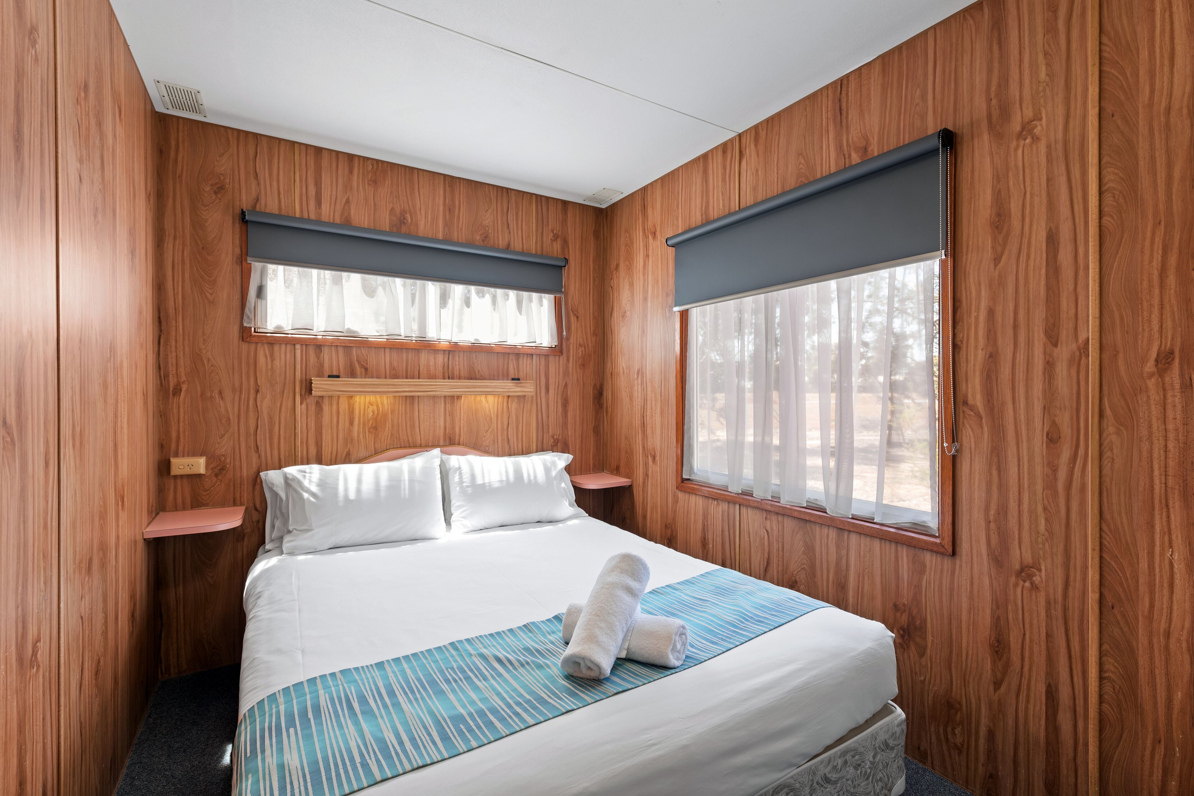 A wooden-paneled room with a bed, two bedside tables, and large windows with curtains.