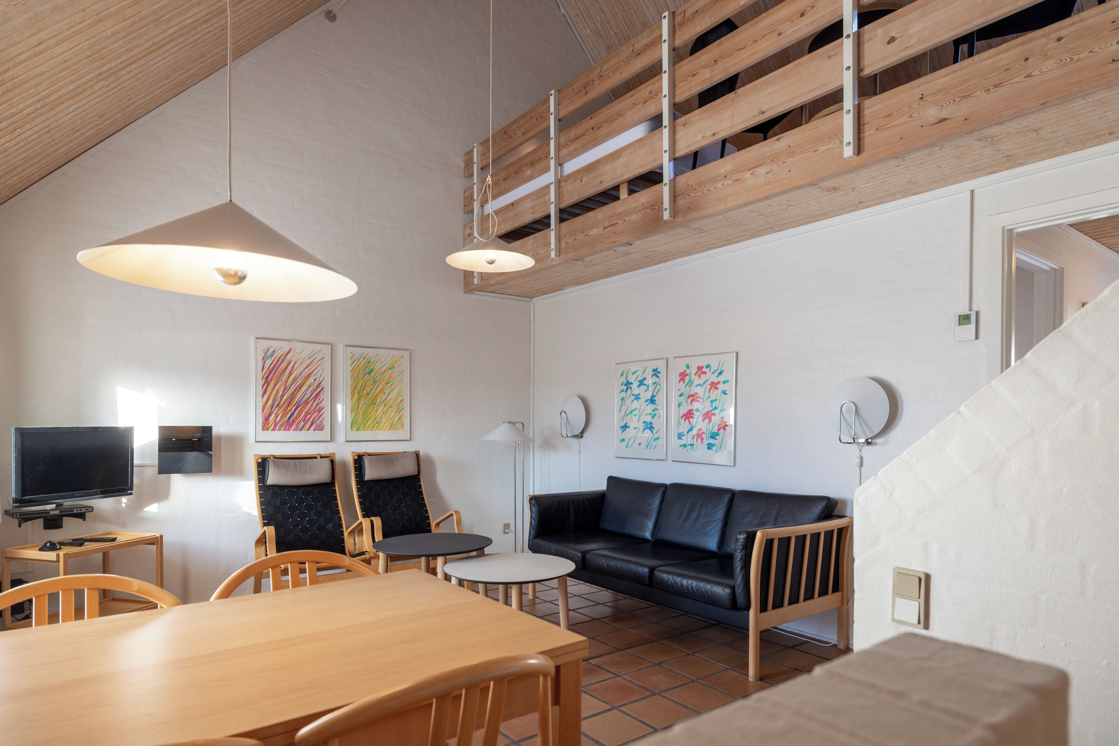 A spacious living area with a wooden dining table, chairs, a black leather sofa, and artwork on the walls.