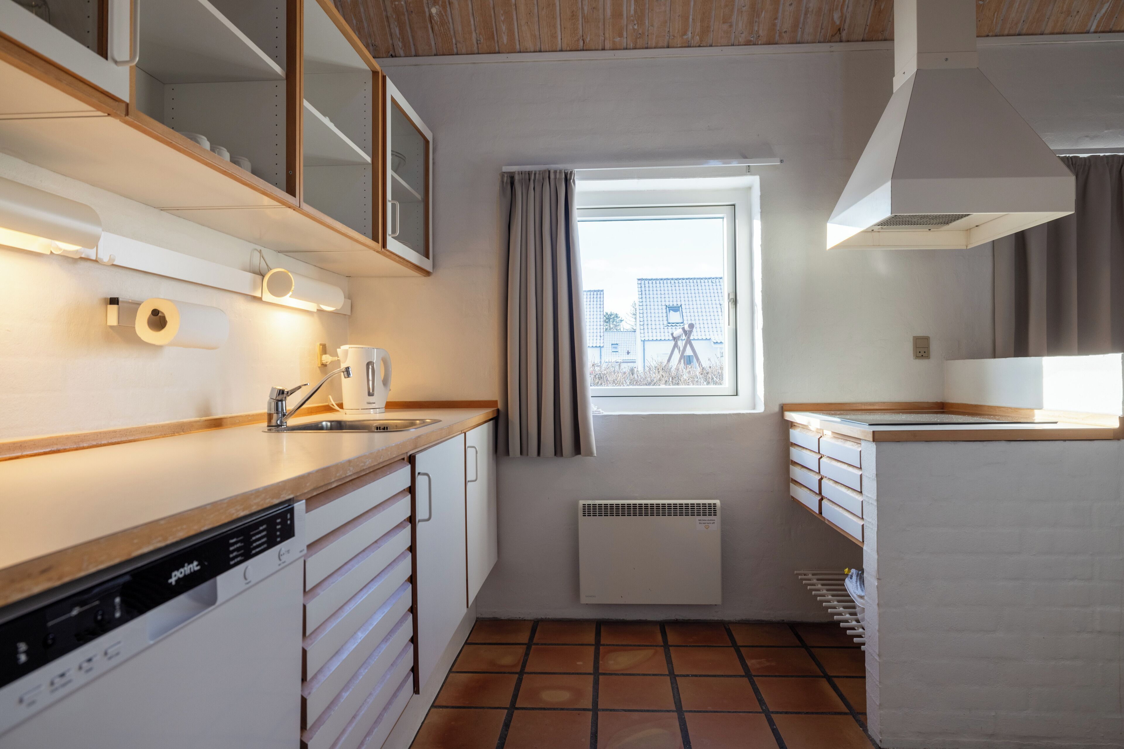 Une cuisine compacte équipée d’un lave-vaisselle, d’une fenêtre avec des rideaux, d’un radiateur et d’un mur en briques blanches.