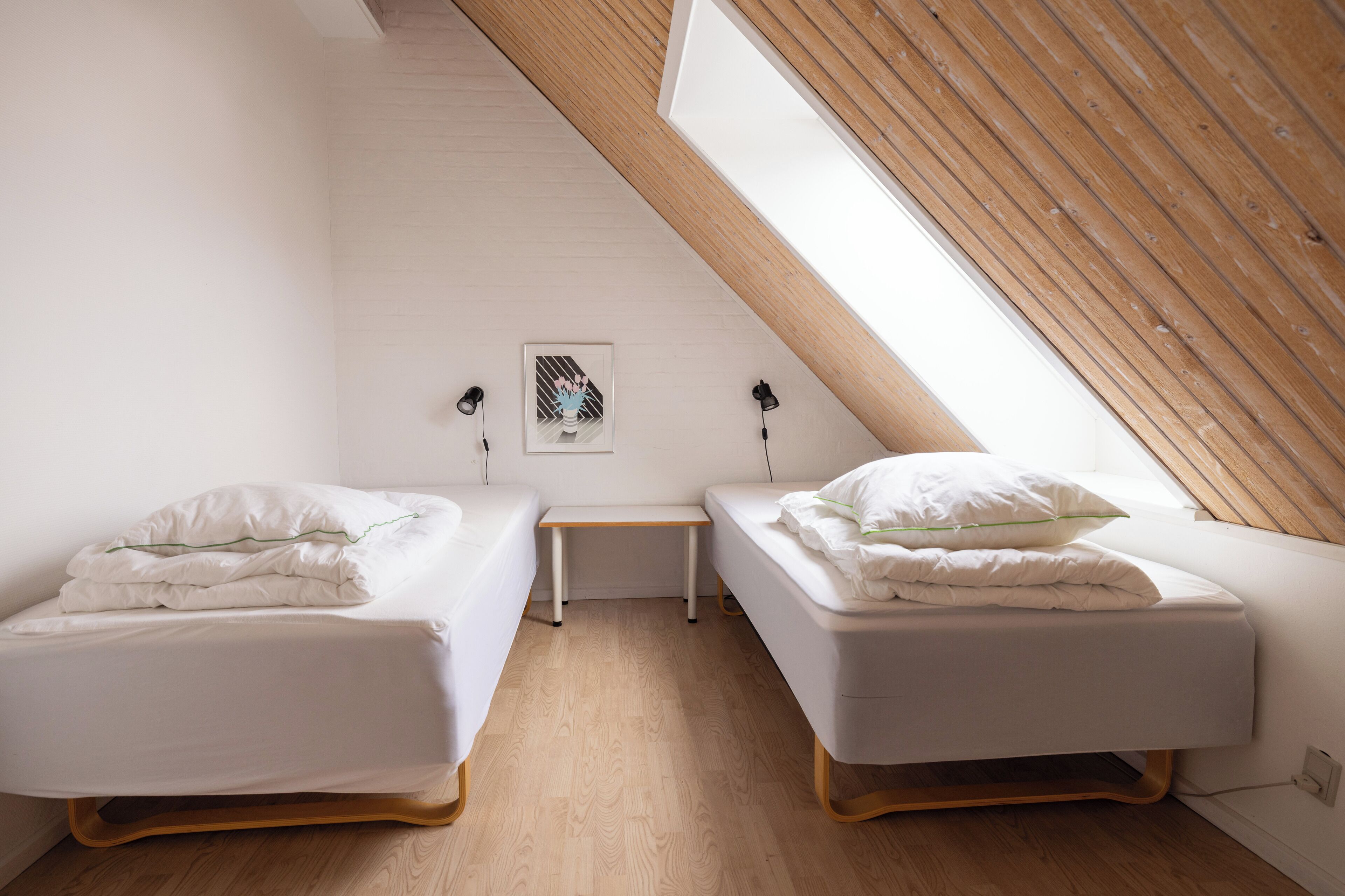 A small, well-lit room with two single beds, a small table, and a sloped ceiling with wooden beams.