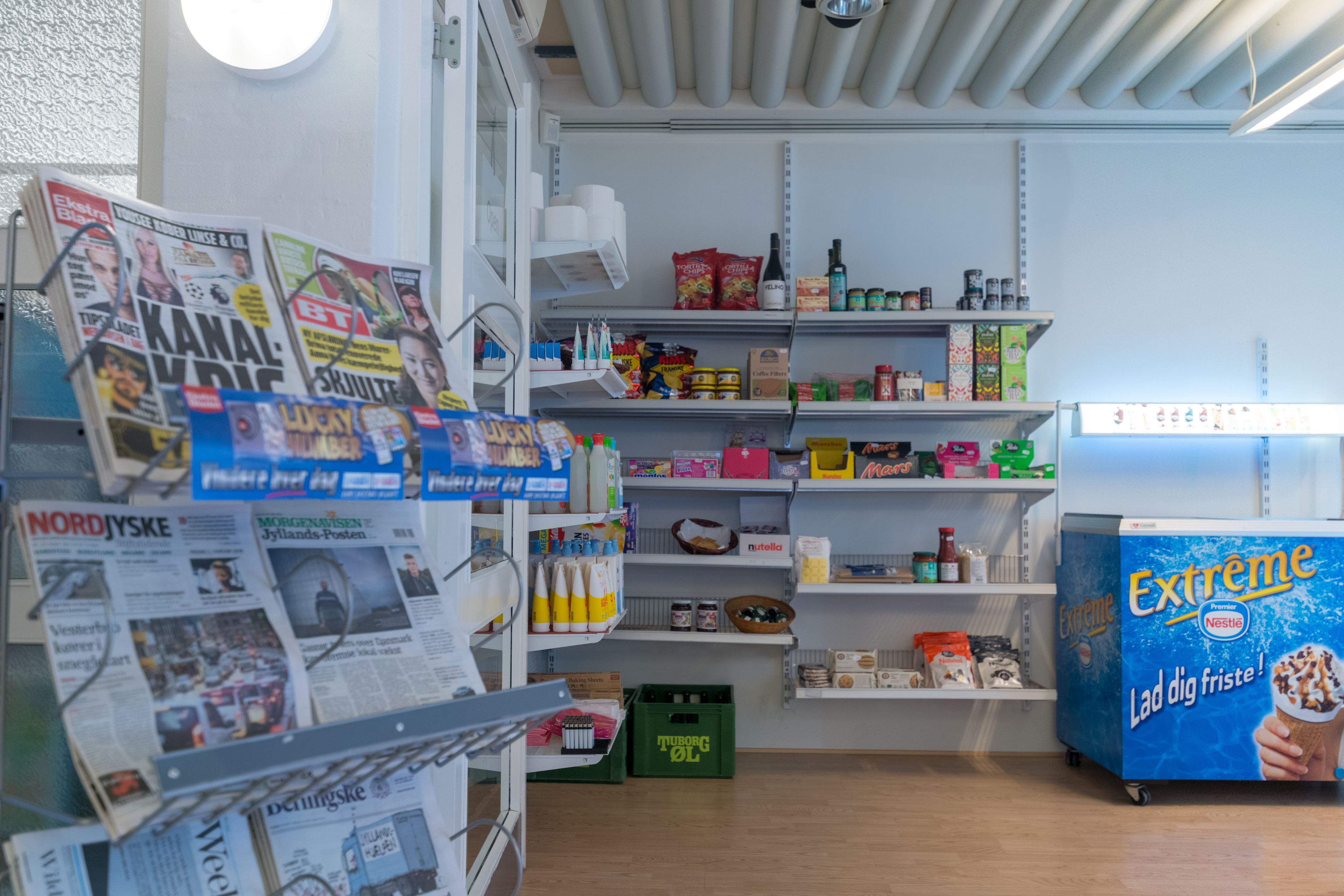 A small, well-organized room with shelves stocked with snacks and beverages, a refrigerator with a Nestle ice cream display, and a wall with newspapers and magazines.