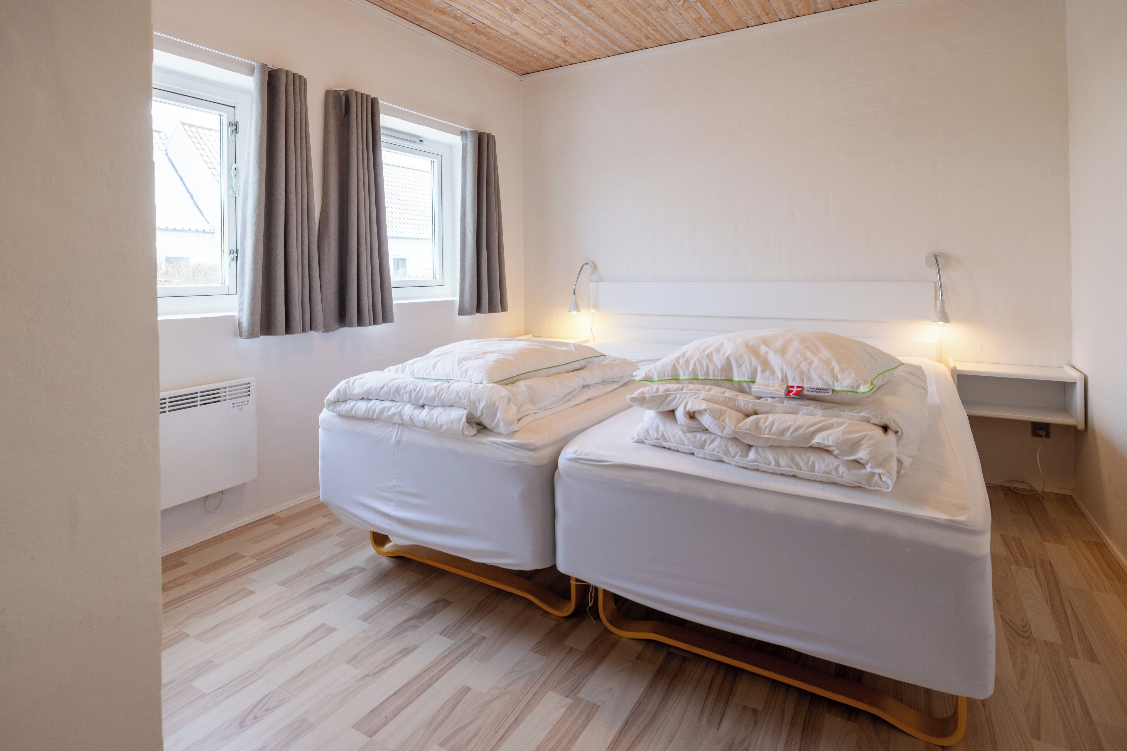 A double bed with white bedding, a wooden floor, a window with curtains, and a wall-mounted nightstand with a lamp.