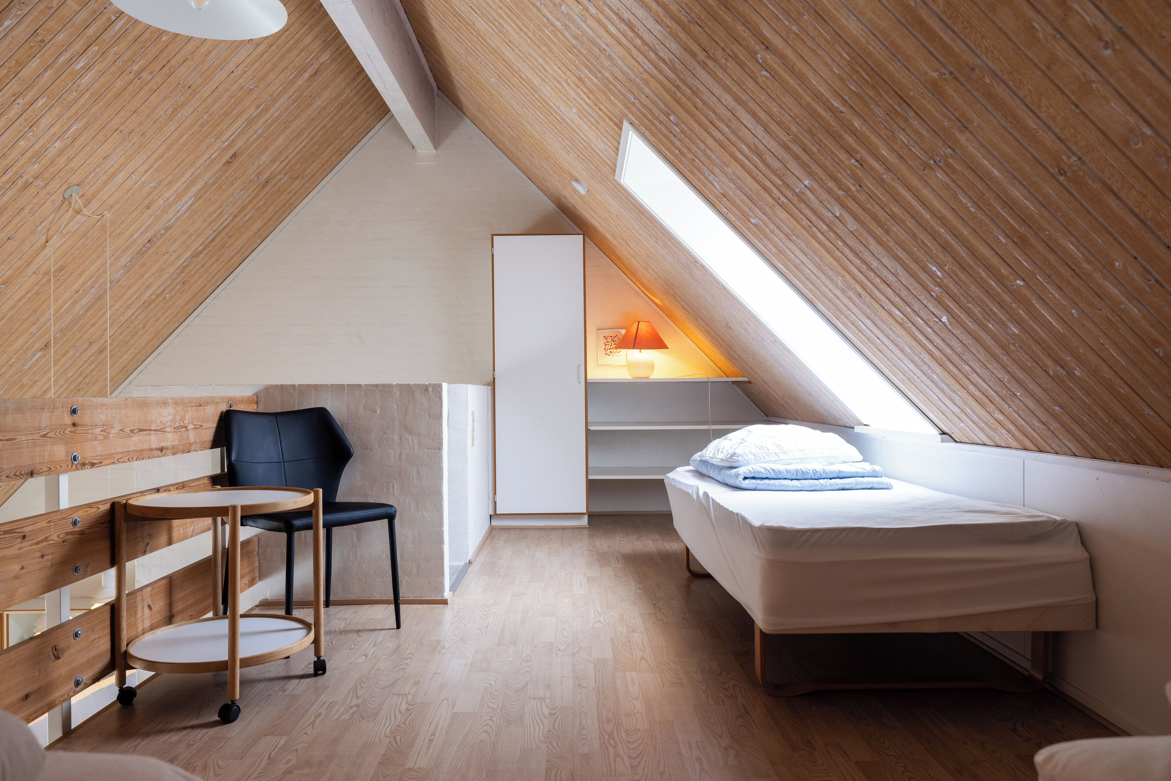 Een compacte, moderne slaapkamer op zolder met een schuine kap, een enkel bed, een stoel, een klein tafeltje en een bureau.