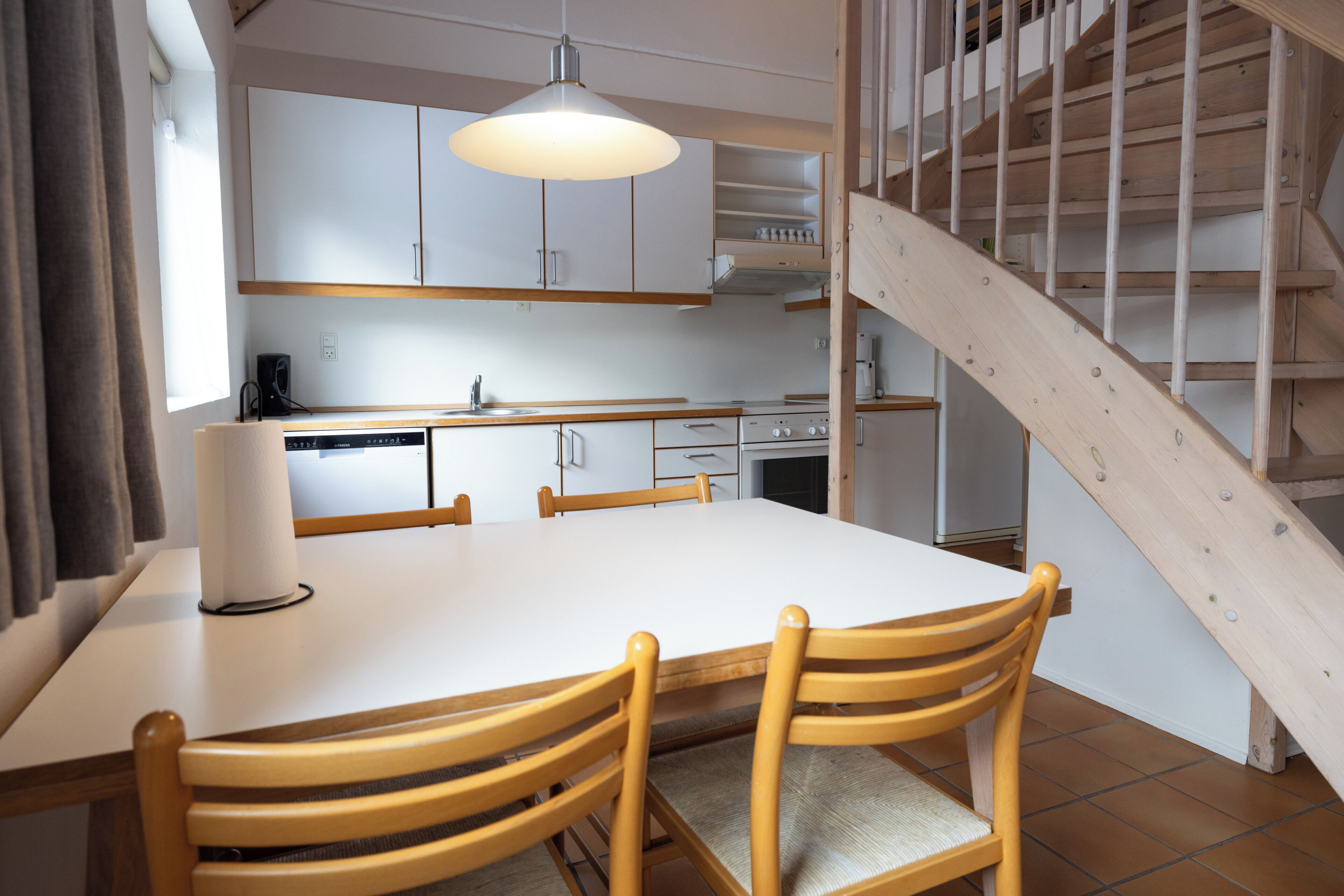A modern kitchen with white cabinets, a wooden dining table, and chairs. There is a staircase with wooden railings leading to an upper level.