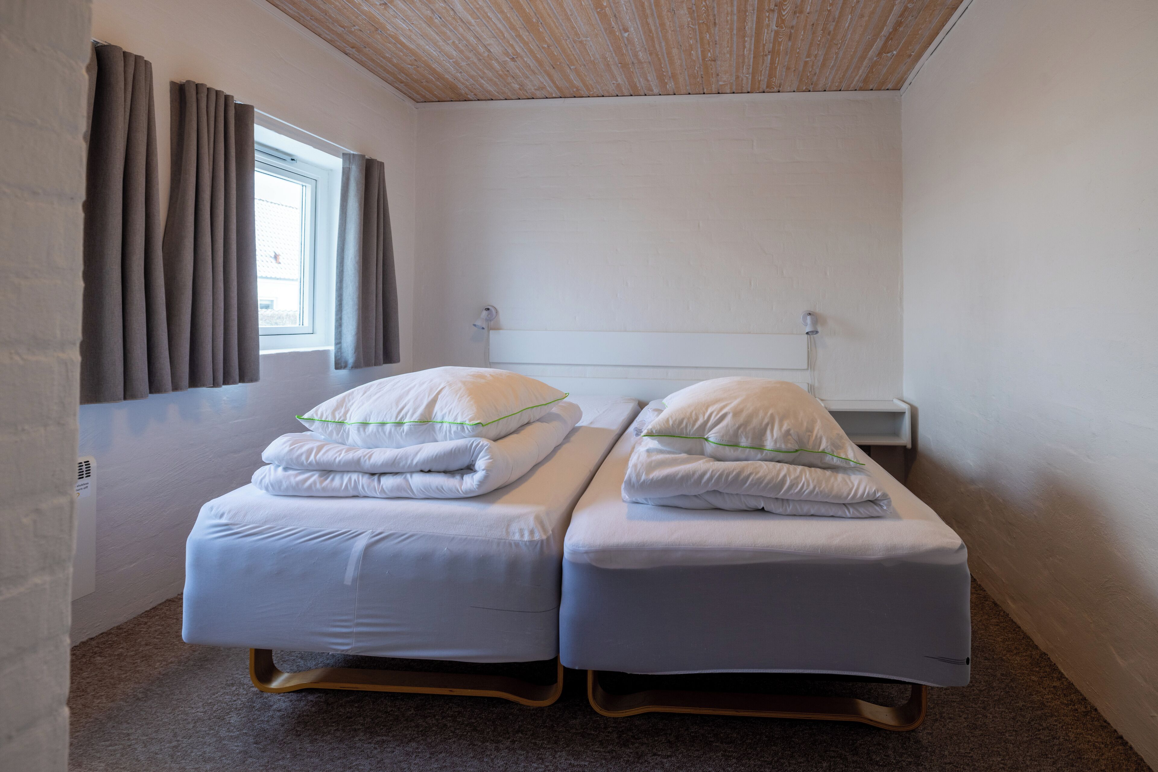 Una habitación con dos camas individuales, una ventana con cortinas, un muro de ladrillo blanco y un techo de madera.
