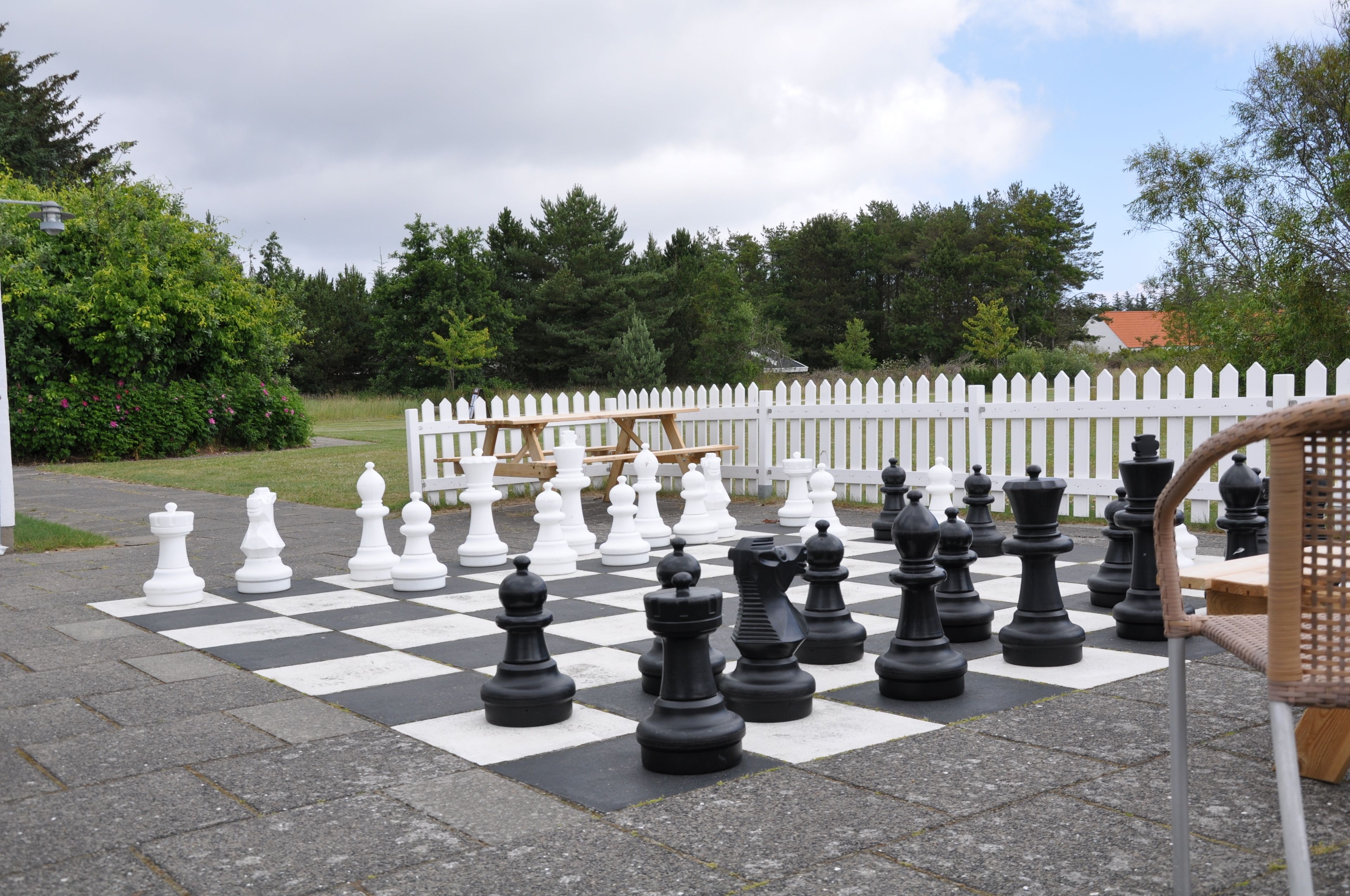 Un grand jeu d’échecs en plein air, installé sur une surface pavée, avec une clôture blanche en bois en arrière-plan.