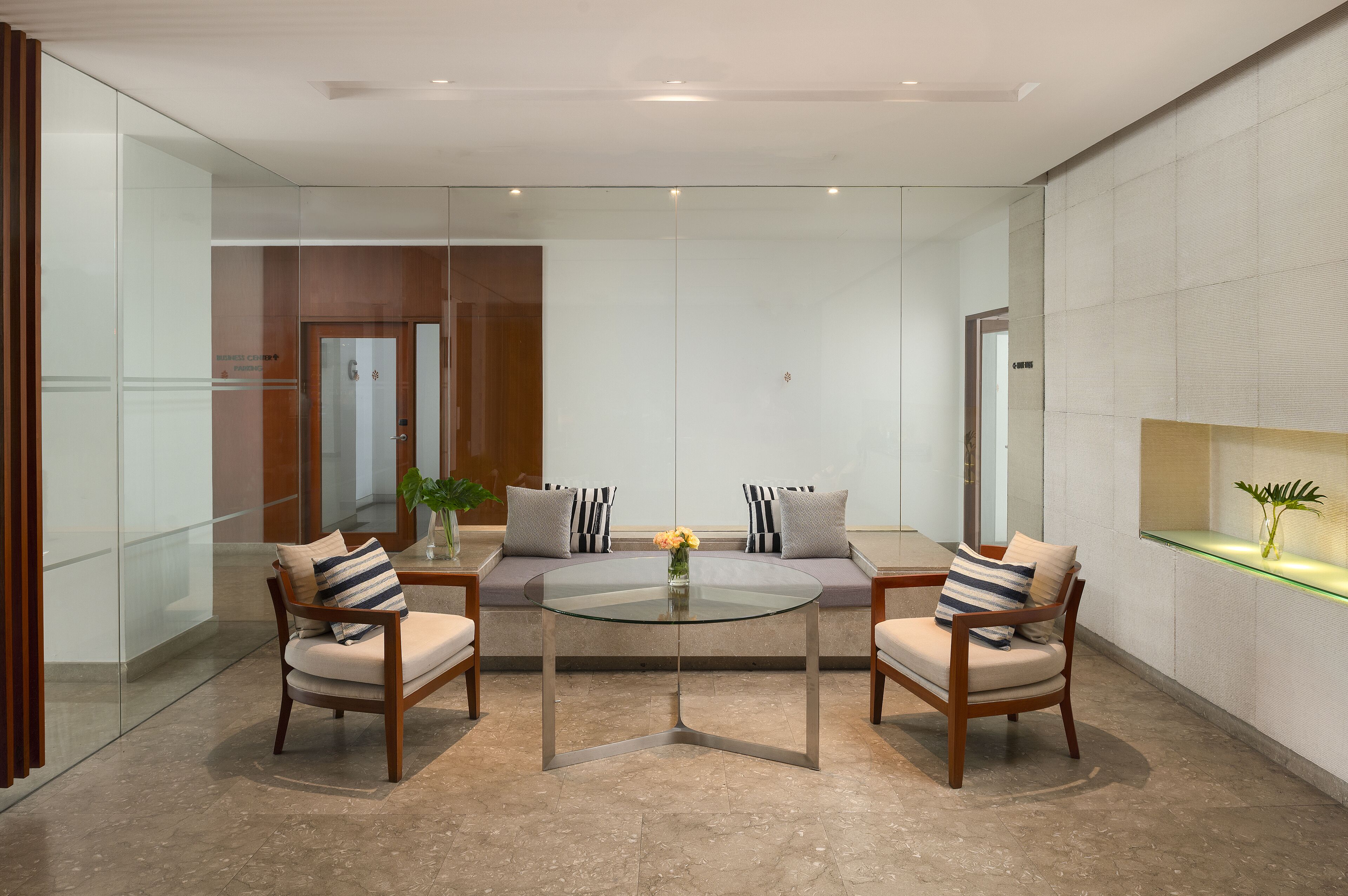 A modern lobby with a glass coffee table, two armchairs, a sofa, and a small plant.