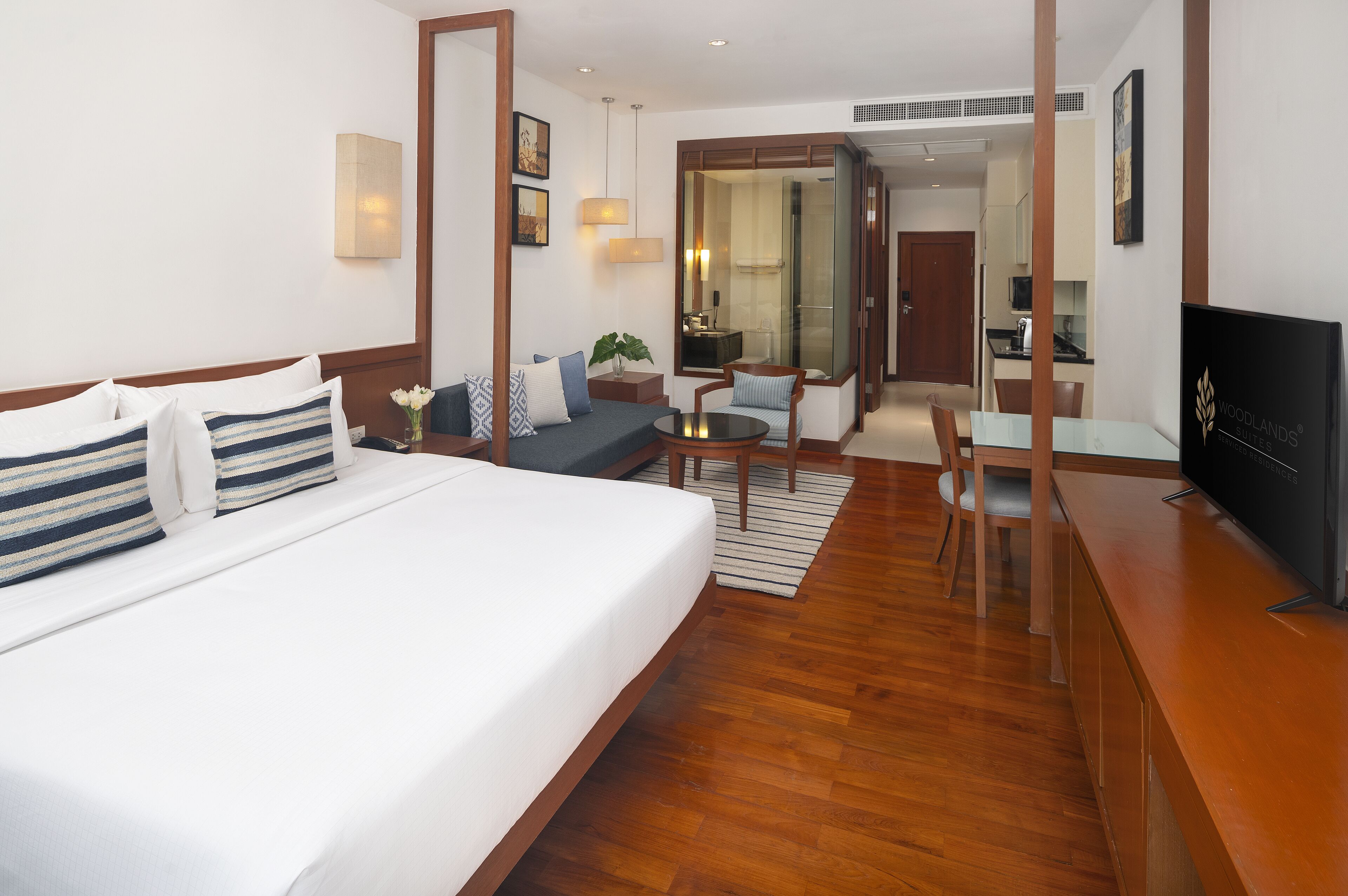 A hotel room with a large bed, a desk, a chair, a TV, and a view of the kitchen and bathroom.