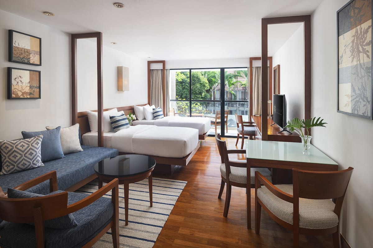 Een moderne hotelkamer met een bank, twee bedden, een eettafel en een groot raam met uitzicht op groen.