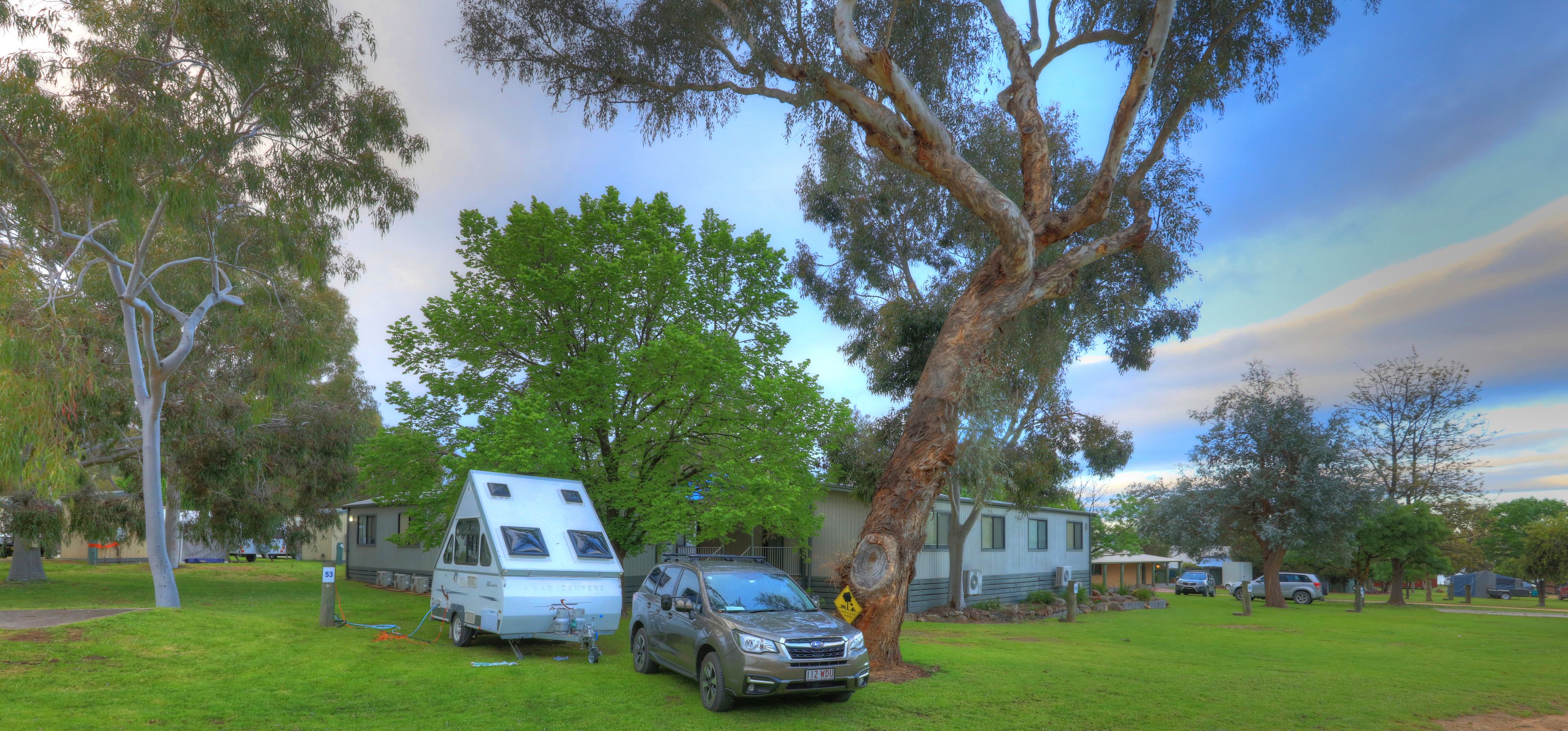 Un camping con una caravana y un coche aparcados en una zona verde, rodeado de árboles y un edificio.