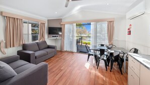 A modern living room with a sofa, dining table, and chairs. There is a ceiling fan, a flat-screen TV, and a fire extinguisher on the wall. - Lake Hume Holiday Park (Bowna)