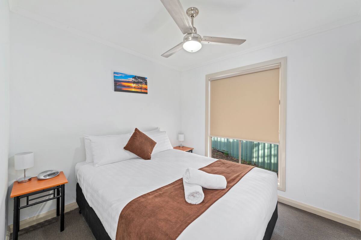Una habitación de hotel con cama, ventilador de techo, mesita de noche con lámpara, un cuadro en la pared y una ventana con persianas.