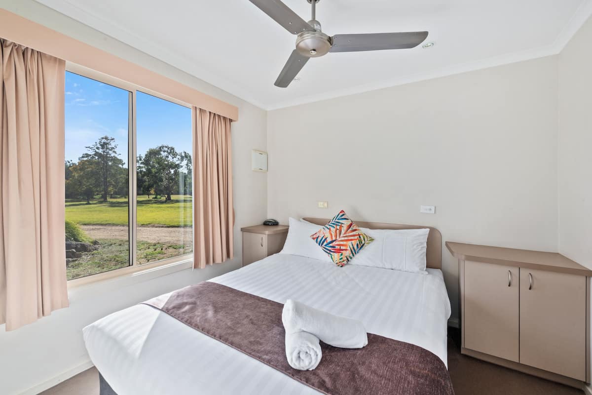 Una habitación de hotel con una cama grande, un ventilador de techo, una mesita de noche, un armario y una ventana con cortinas.