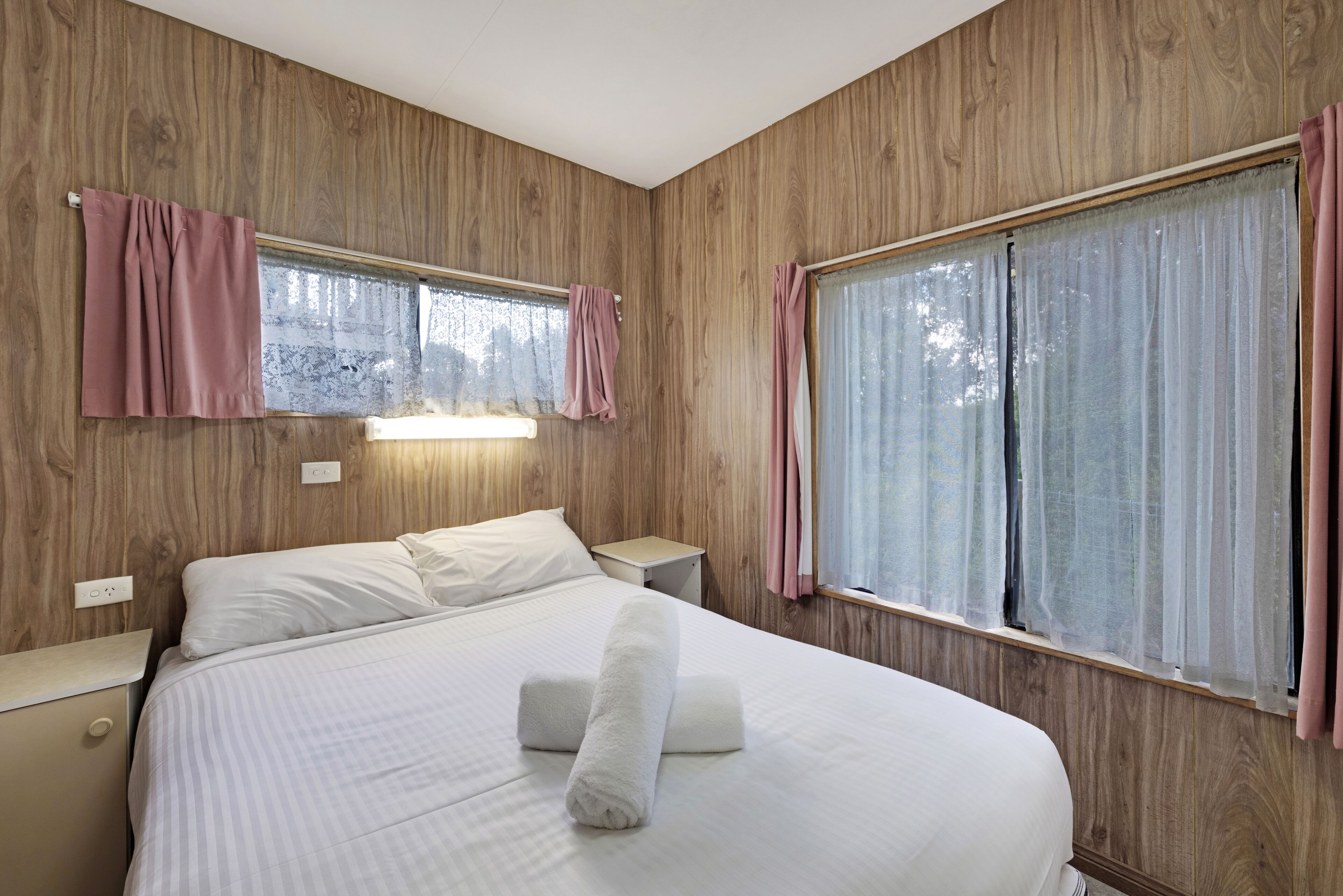 A hotel room with a wooden paneled wall, a large bed with white bedding, two bedside tables, and a window with sheer curtains.