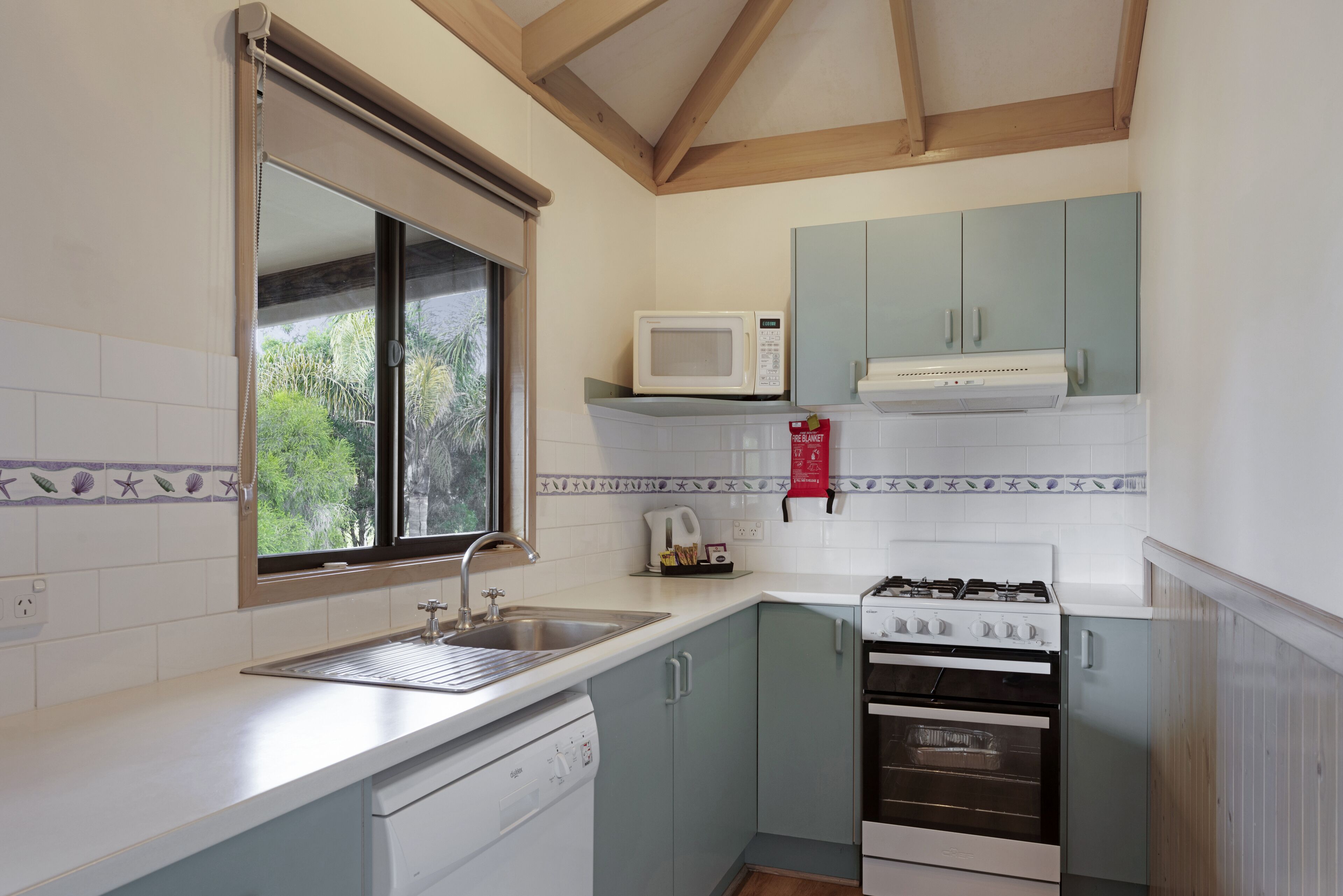 A compact kitchen with a dishwasher, stove, microwave, and a window with a view of greenery.