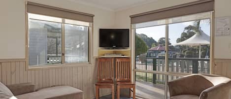 Una habitación con televisión sobre un soporte de madera, dos sillas de madera, un sofá y ventanas grandes con persianas.