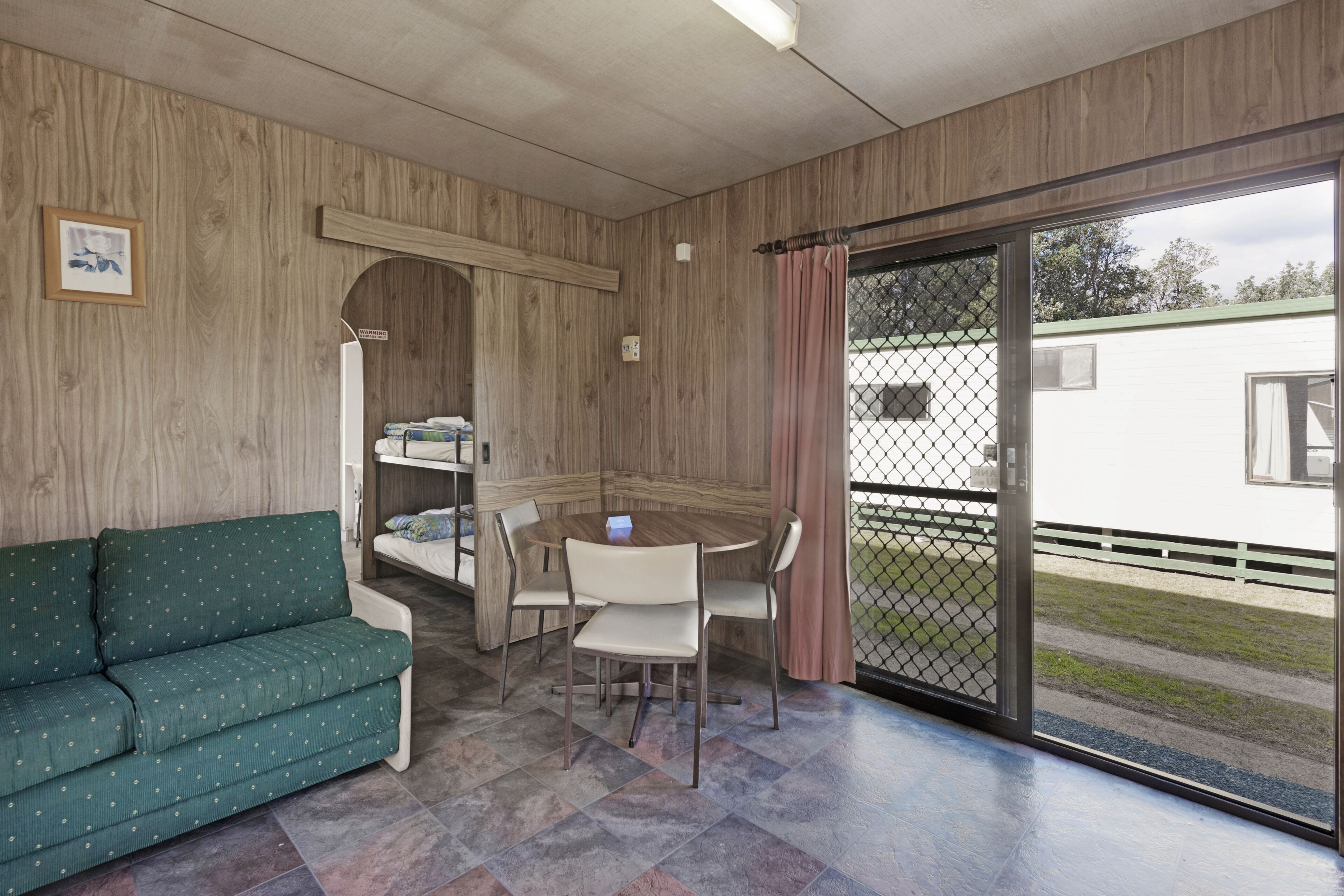 A room with a wooden wall, a green sofa, a dining table with chairs, and a sliding glass door leading outside.