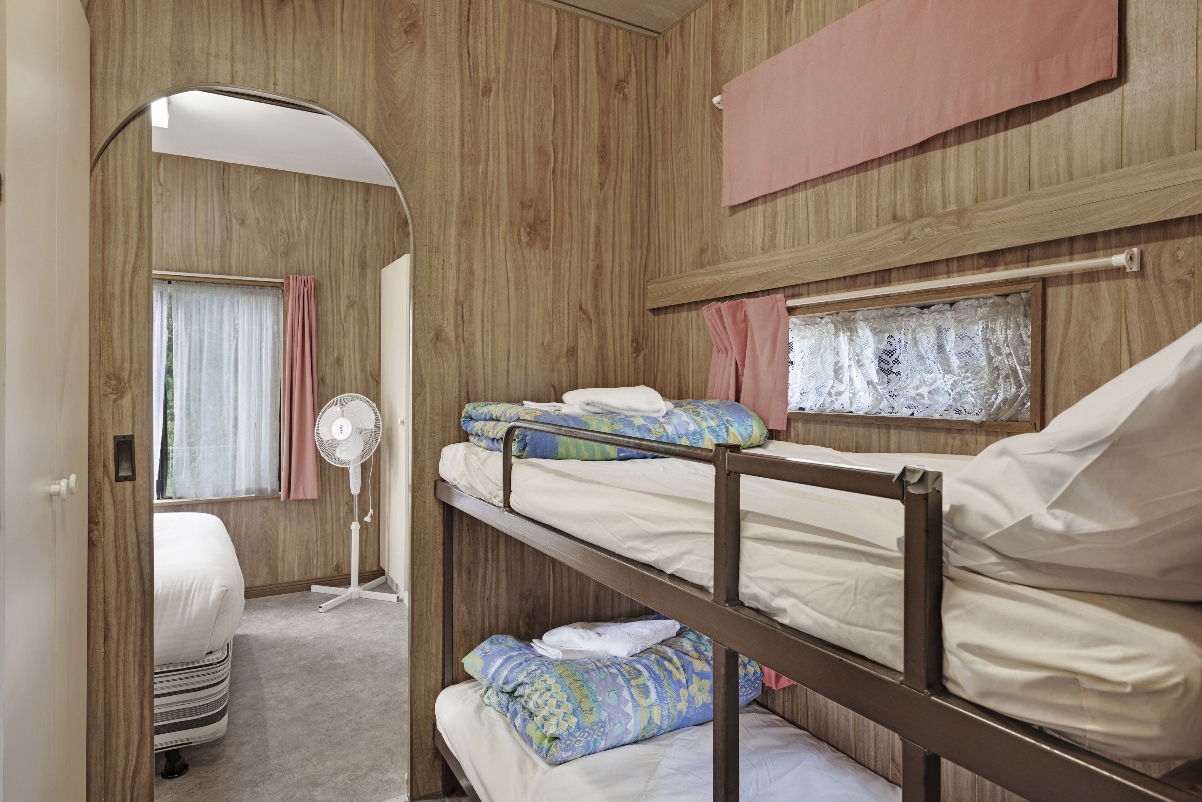 Une chambre avec un lit superposé, des murs en bois, un ventilateur sur pied et une fenêtre avec des rideaux.