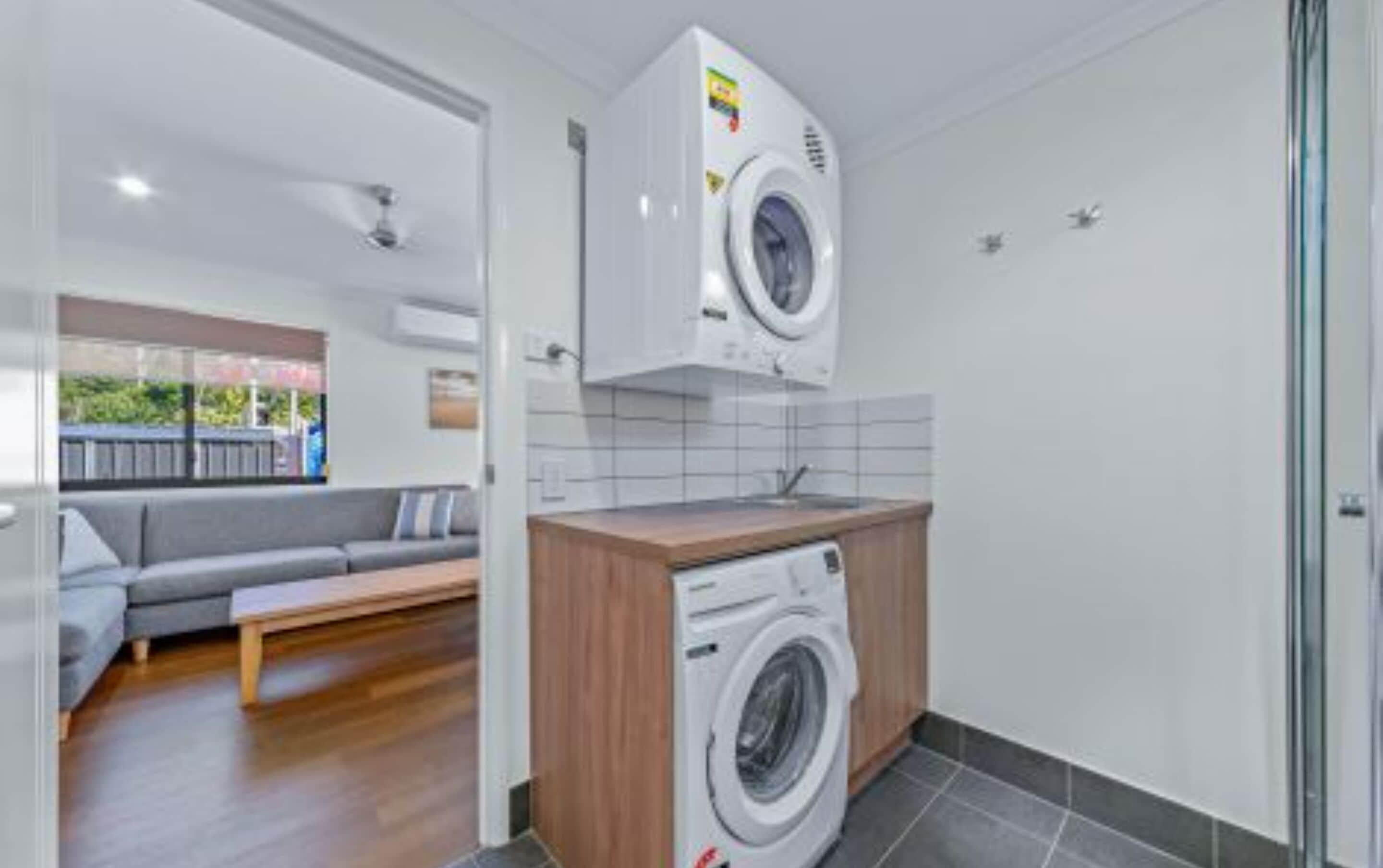 A compact laundry room with a washing machine and dryer, a wooden counter, and a grey sofa in the adjacent living area.