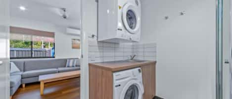 A compact laundry room with a washing machine and dryer, a wooden counter, and a grey sofa in the adjacent living area.