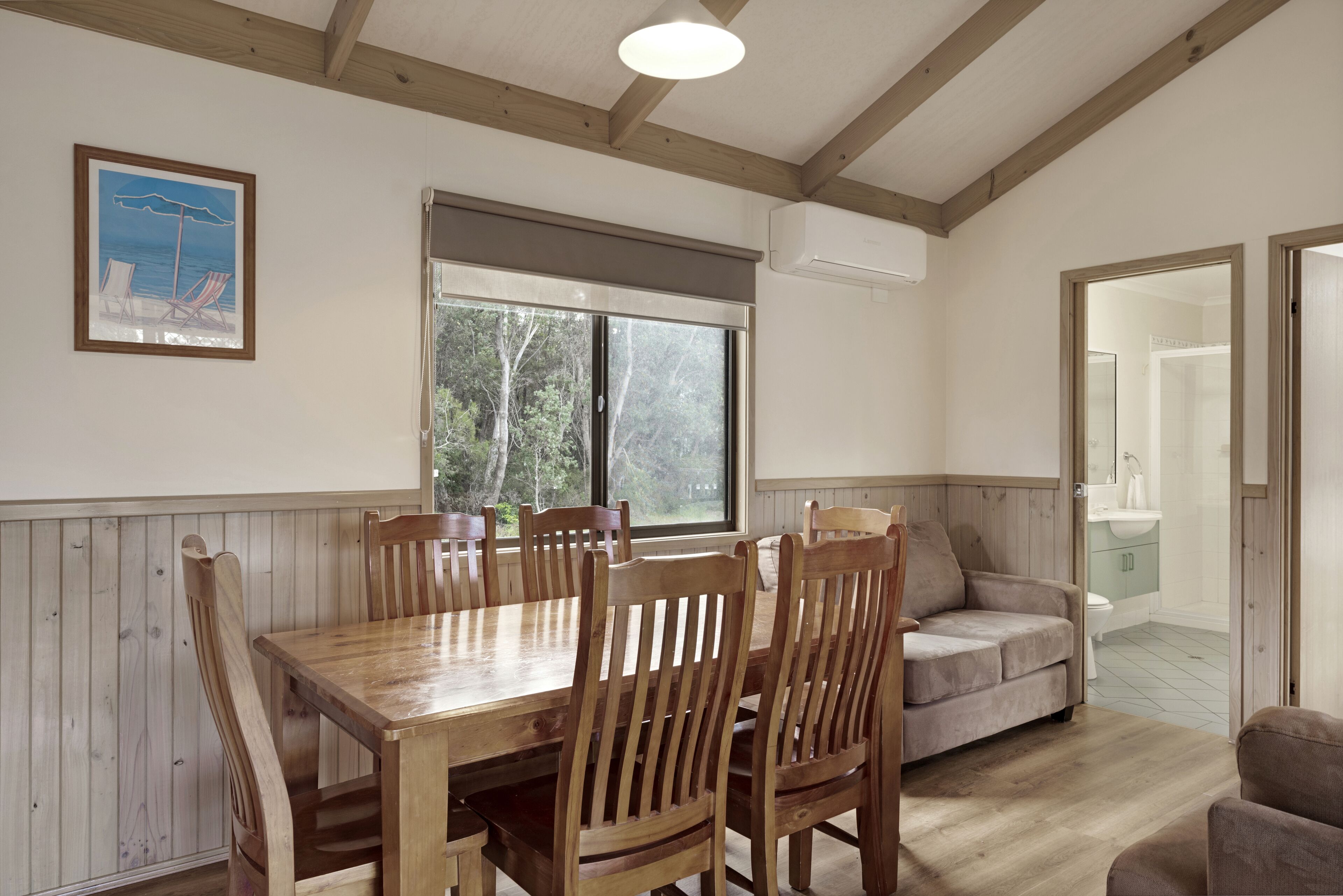 A dining area with a wooden table and chairs, a window with a view of trees, a framed picture on the wall, and a sofa in the background.