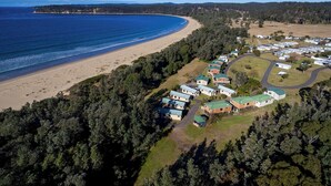 An aerial view of a coastal area with a beach, dense forest, and a row of mobile homes with green roofs. - Discovery Parks - Eden (Boydtown)