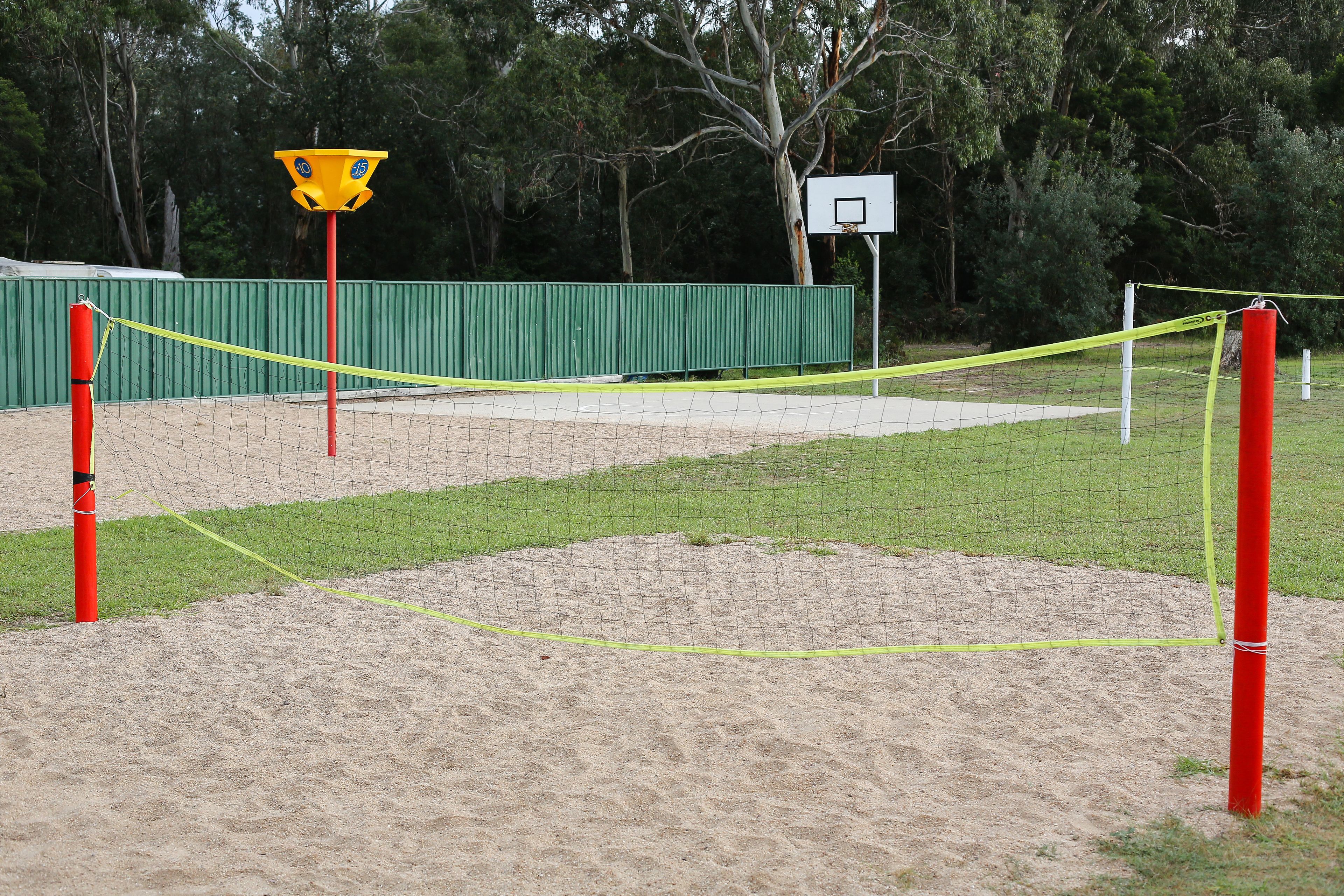A sand volleyball court with a net, sand, and a basketball hoop in the background.
