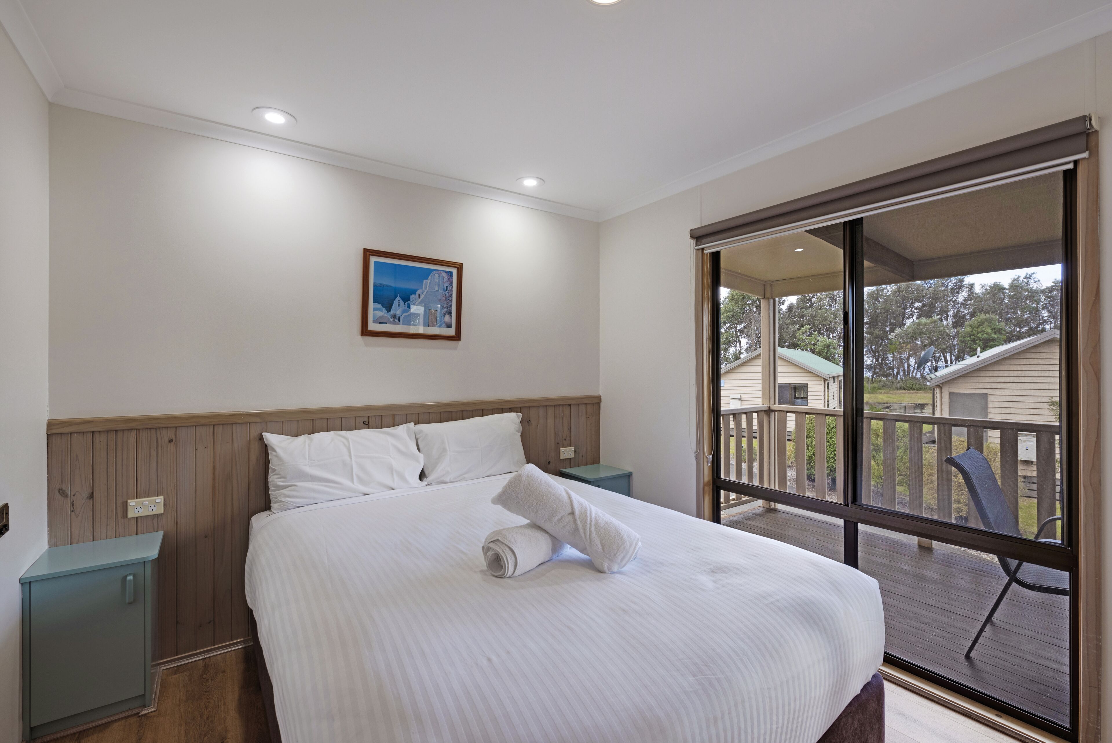 A bedroom with a large bed, a nightstand, a wooden headboard, a painting on the wall, and a balcony with a view of other buildings.