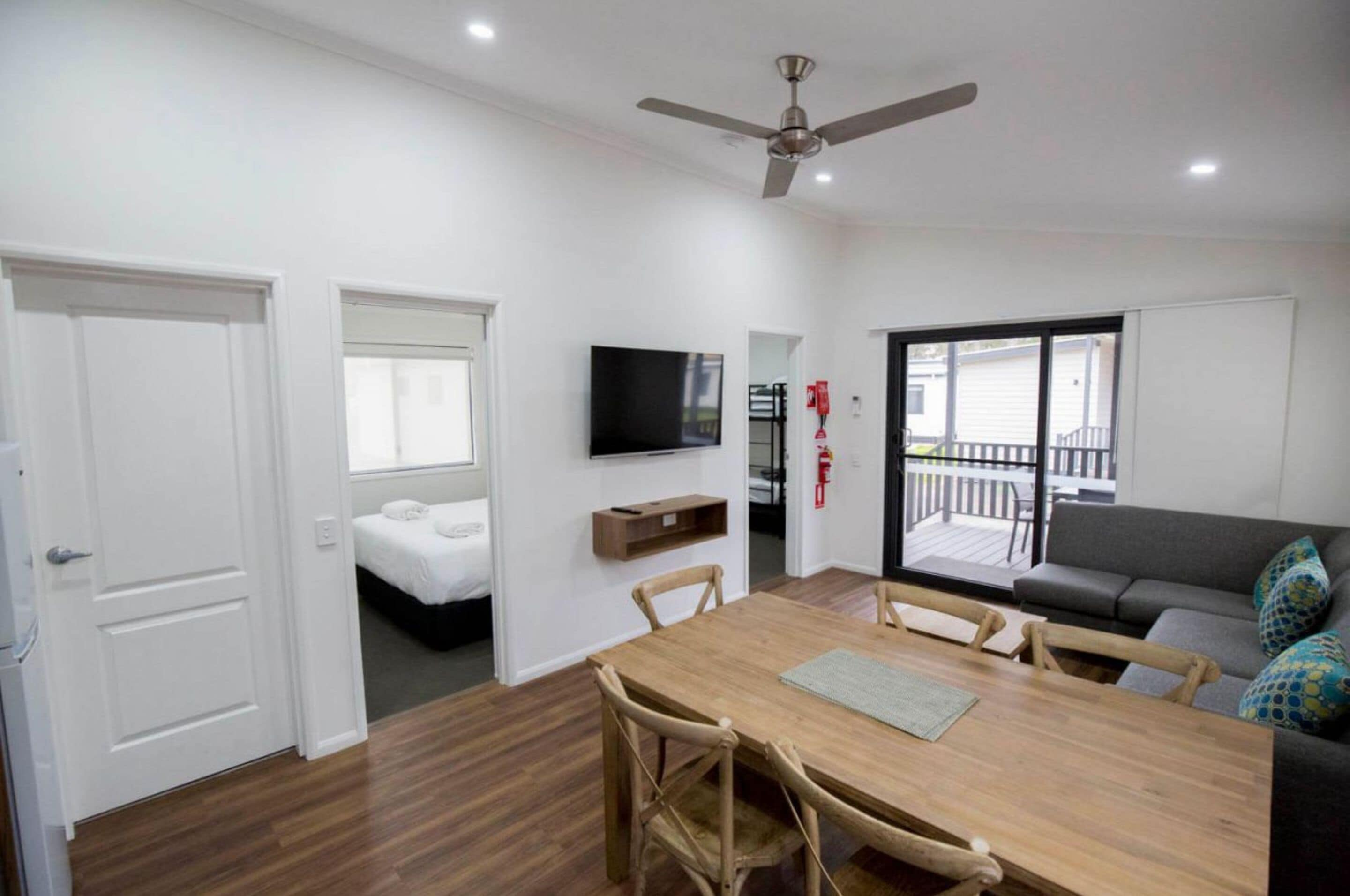 A modern living room with a wooden dining table, a sofa, a TV mounted on the wall, and a balcony with a railing.