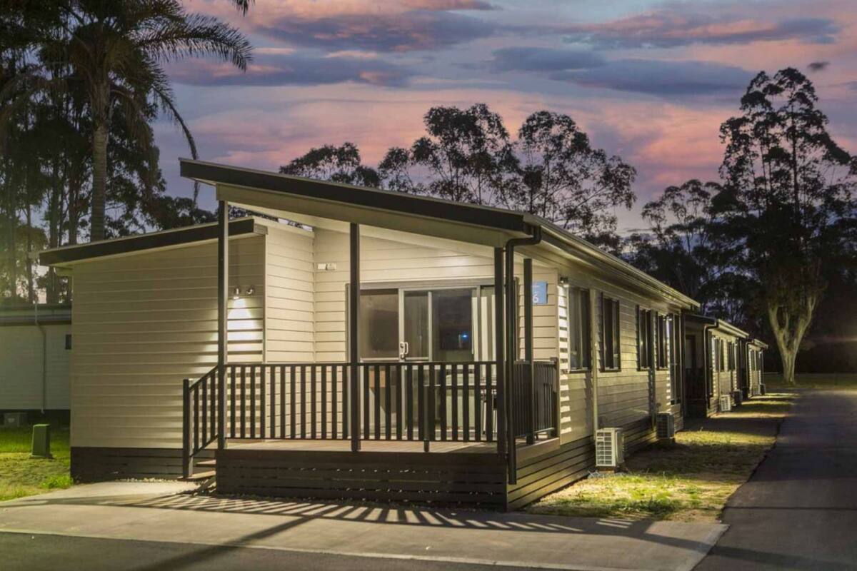 A single-story mobile home with a porch and a covered entryway, surrounded by trees and a clear sky.