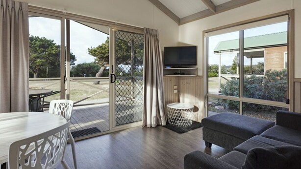 A modern living room with a sofa, a TV, and a dining area with a table and chairs. - Discovery Parks - Eden (Boydtown)